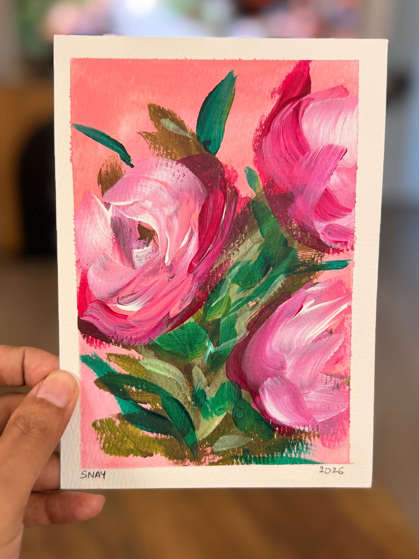 A hand holds a small art print or card featuring an expressive painting of pink flowers with green leaves against a pink background, with a blurred indoor room behind it.
