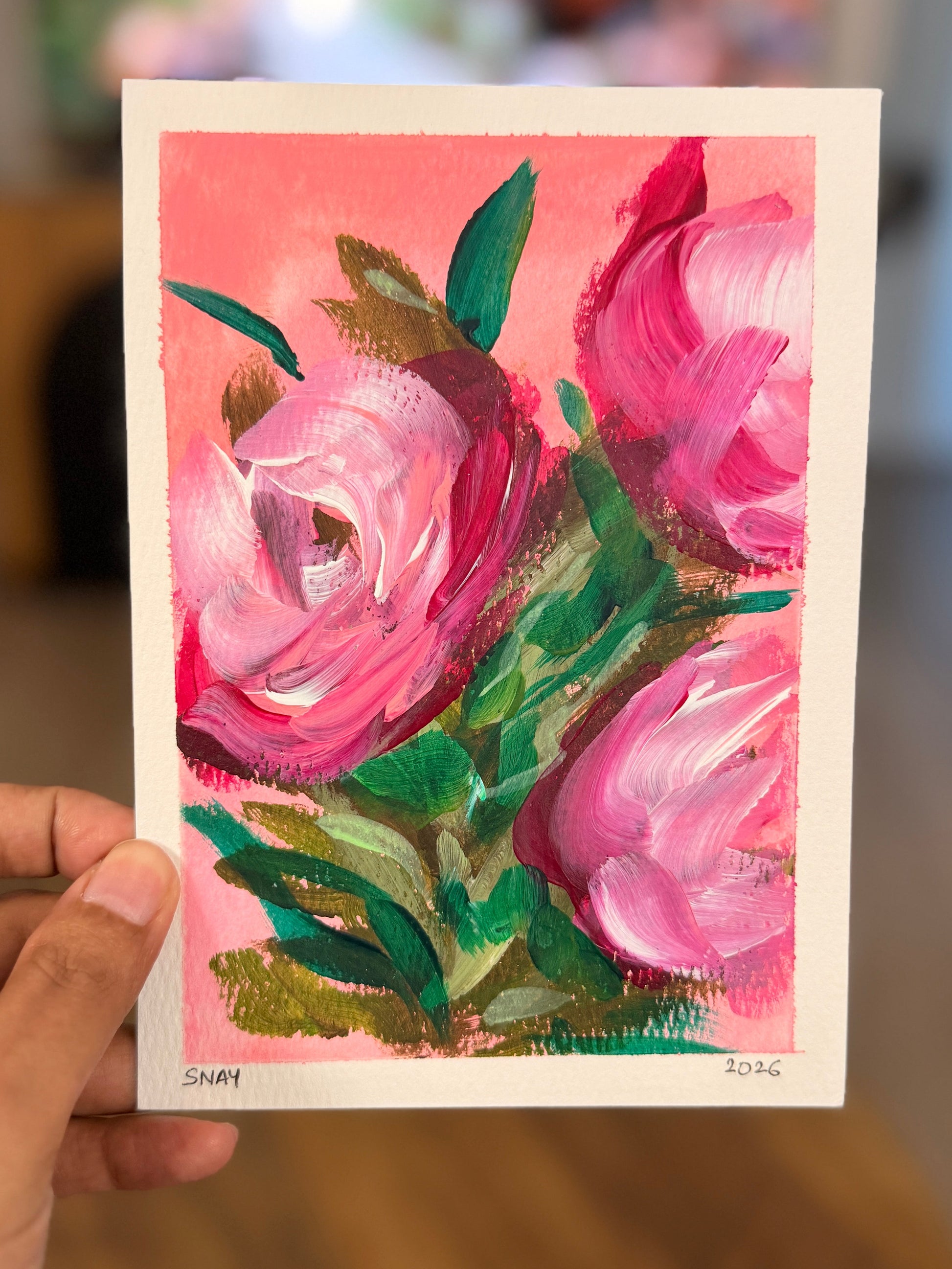A hand holds a small art print or card featuring an expressive painting of pink flowers with green leaves against a pink background, with a blurred indoor room behind it.