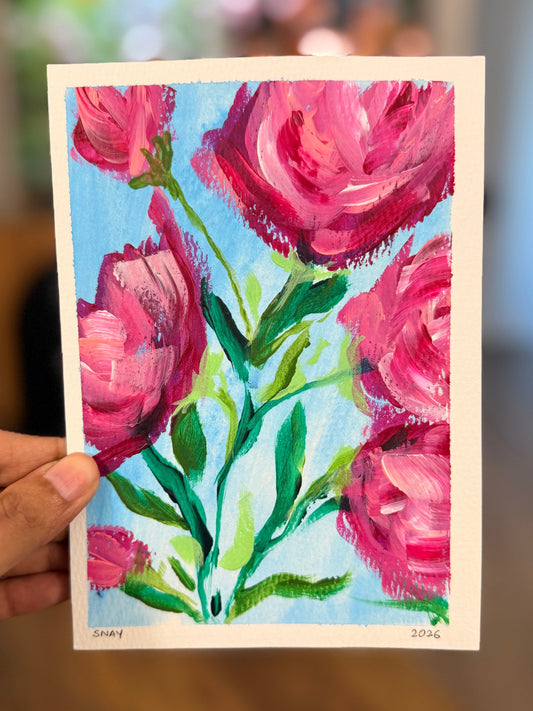A hand holds a square acrylic painting of pink roses with green stems against a light blue background, with a blurred indoor room behind it.