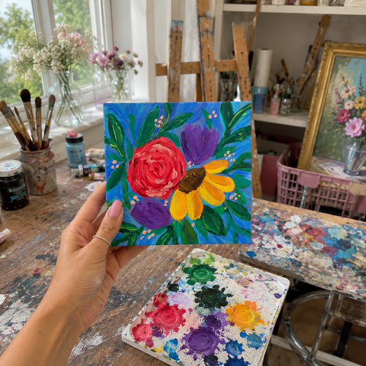 Hand holding a floral painting in an artist's studio with art supplies and other paintings.