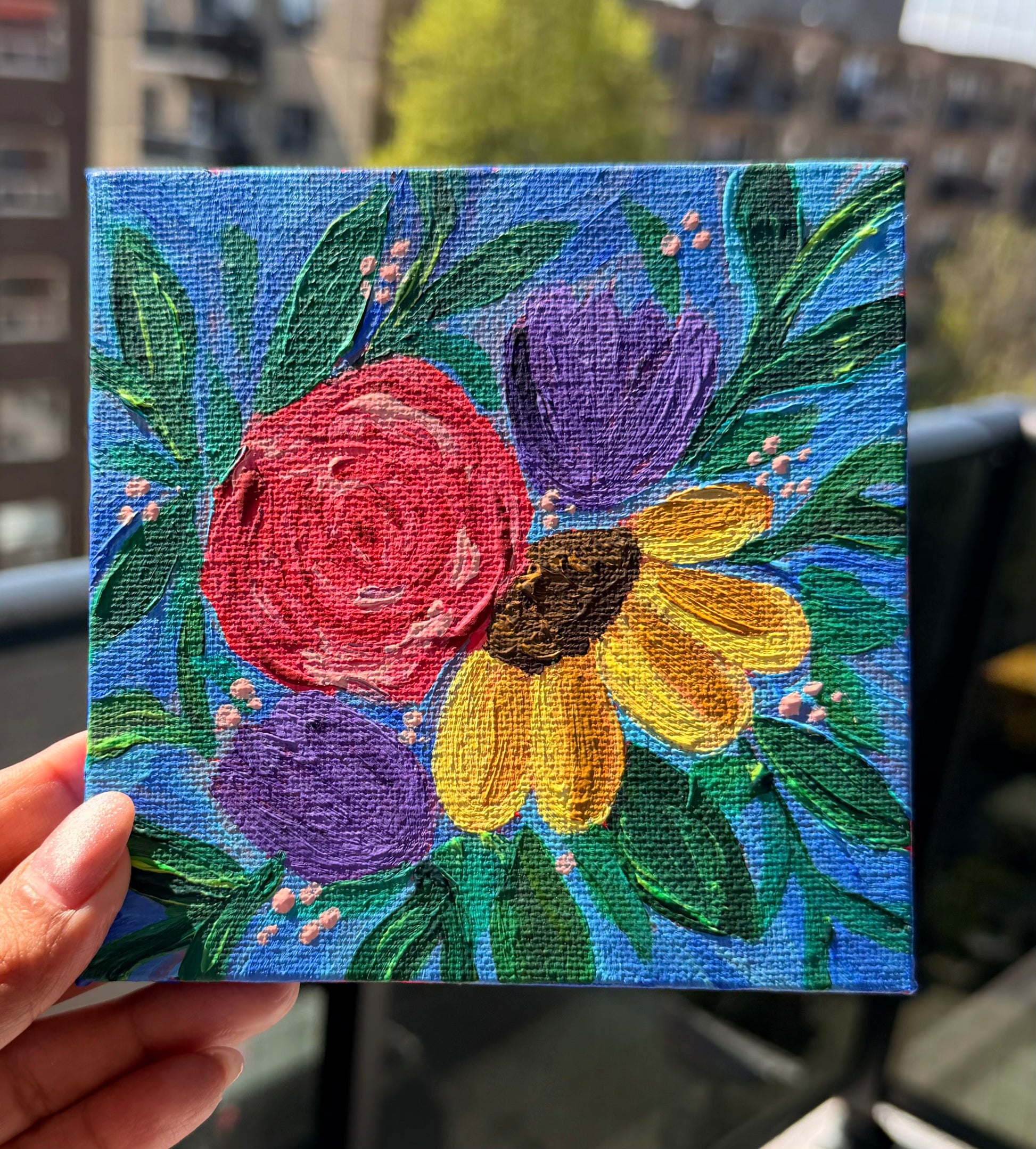 Small floral painting held by a hand with a blurred urban background